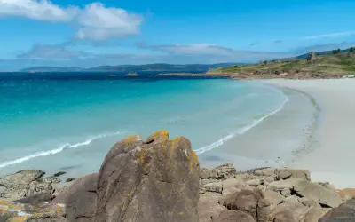 Aguas turquesas y arena blanca en una de las hermosas playas de la Costa da Morte. Rocas cubiertas de líquenes en primer plano.
