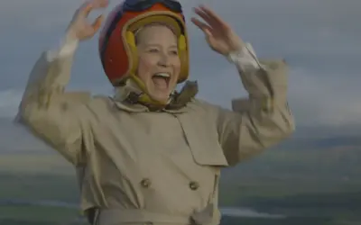 Glad Trine Dyrholm i pilotmössa och trenchcoat vinkar glatt. En scen från filmer med Trine Dyrholm.