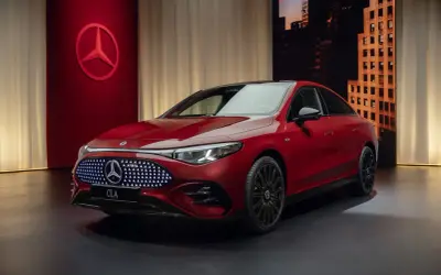 Elegancki, czerwony nowy Mercedes CLA z podświetlanym grillem i czarnymi felgami stoi na ciemnym tle z miejskim krajobrazem w tle.