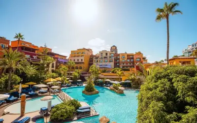 Sonnige Poolanlage mit bunten Gebäuden und Palmen im Riu Palace Teneriffa. Entspannung pur unter blauem Himmel.