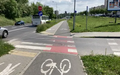 Pasy na ścieżce rowerowej i pasy dla pieszych przecinają drogę. Autobus i samochody czekają na zielone światło.