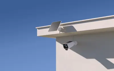En eufycam 2 pro test visar en vit övervakningskamera med solpanel monterad på en vit vägg under en blå himmel.