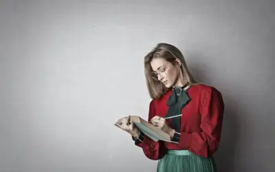 Młoda kobieta w okularach i czerwonej bluzce z zieloną kokardą pochyla się nad książką, notując coś ołówkiem. To inspiracja, jak napisać esej historyczny.