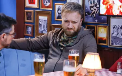 Tatusiowie serial obsada: Mężczyzna z brodą i w bluzie siedzi przy stole z piwem, w tle ściana z ramkami.