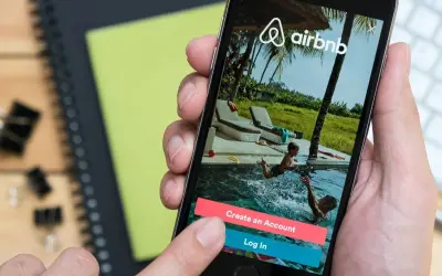 Dłoń trzyma smartfon z aplikacją Airbnb. Widok na basen z dziećmi. Zastanawiasz się airbnb co to? To platforma do rezerwacji noclegów.