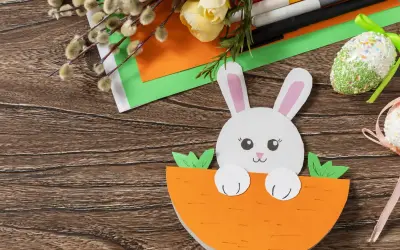 Uroczy króliczek z papieru kolorowego wychyla się z marchewki. Obok pisanki z papieru kolorowego, bazie i flamastry.