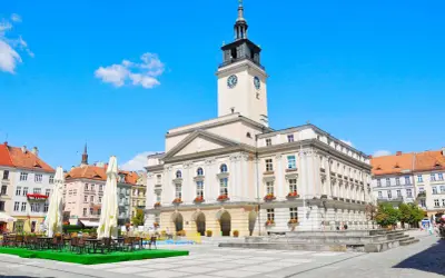 Rynek w mieście nad Prosną. Ratusz z wieżą zegarową góruje nad placem, gdzie stoją stoliki kawiarniane.
