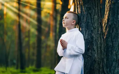 Chłopiec w albie modli się w lesie, promienie słońca przebijają się przez drzewa. Piękne sesje komunijne w plenerze.