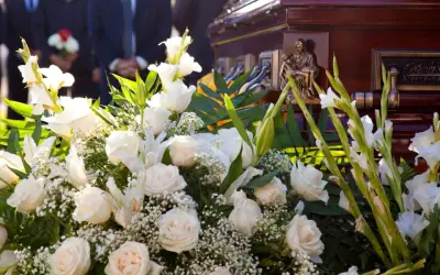 Elegancki wieniec pogrzebowy z białych róż i lilii, ozdobiony zielenią, przy trumnie. Cena 200 zł.