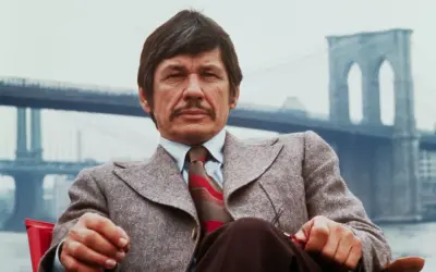 Charles Bronson w garniturze, z wąsami, siedzi przed mostem Brooklińskim. Ikona kina akcji, znany z wielu filmów.