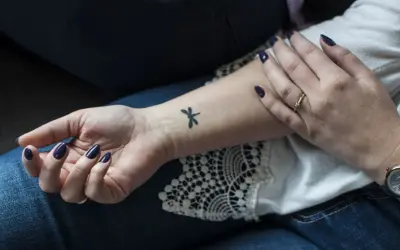 Delikatne, czarne tatuaze male na nadgarstku, w tym ważka. Dłoń ozdobiona ciemnym lakierem i pierścionkiem.