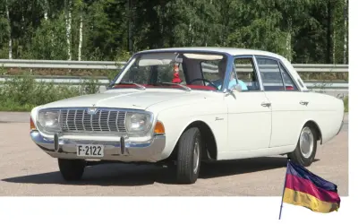 Vit Ford Taunus, en klassisk modell av svenska bilar, parkerad utomhus med en tysk flagga i förgrunden.