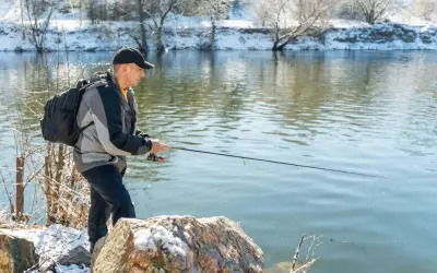 Mann angelt bei Minusgraden am Flussufer, das mit Schnee bedeckt ist. Er trägt eine Jacke und einen Rucksack.