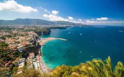Vista panoramica della costa con il **scoglio del cavaliere** che emerge dal mare turchese, spiagge affollate e case aggrappate alla scogliera.