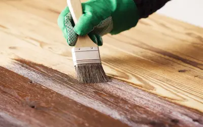 Mano con guante verde barnizando mesa de madera con brocha. El acabado protege y embellece la veta.
