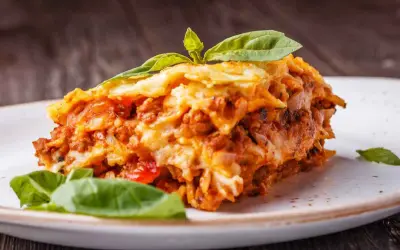Una fetta di lasagna salentina, ricca e saporita, guarnita con basilico fresco, su un piatto bianco.