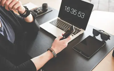 Osoba trzyma iPhone'a, obok leży tablet i laptop z widocznym zegarem.