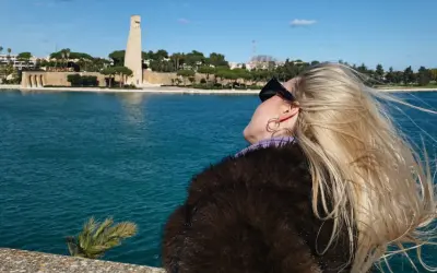 Kobieta z blond włosami podziwia pomnik w Brindisi. W tle błękitne morze i błękitne niebo.