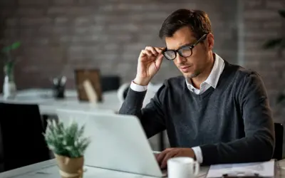 Mężczyzna w okularach, skupiony na laptopie, pracuje przy biurku. W tle widać elementy biurowe, tworząc atmosferę miejsca pracy. Ten sygnalista w pracy analizuje dane.