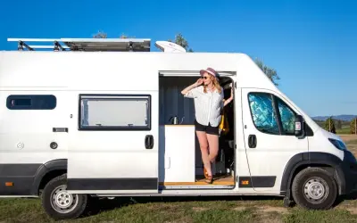 Kobieta w kapeluszu wychodzi z białego busa kampera, ciesząc się słonecznym dniem na tle błękitnego nieba.