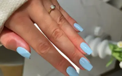 Dłoń z paznokciami w kolorze baby blue, ozdobiona złotym pierścionkiem z brylantem.
