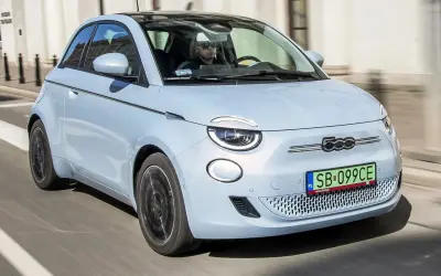 Jasnoniebieski fiat elektryczny 500e z czarnym dachem jedzie ulicą. Kierowca w okularach przeciwsłonecznych prowadzi auto.