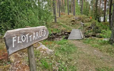 Träskylt med texten "Flottarleden Munkedal" pekar mot en liten träbro över en bäck i en skogsmiljö.