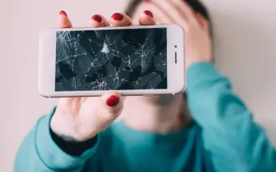 Złamany telefon z pękniętym ekranem, który osoba trzyma przed twarzą, zakrywając ją ręką.