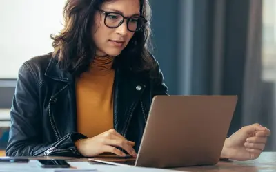 Kobieta w okularach i skórzanej kurtce pracuje na laptopie, szukając informacji, jak sprawdzić lata pracy w ZUS PUE.