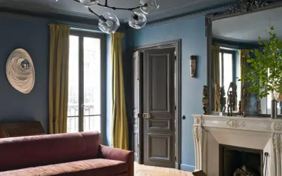 Soggiorno elegante con pareti blu balena Pantone, divano bordeaux, tendaggi ocra e camino in marmo.