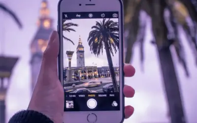 Dłoń trzyma iPhone'a, na ekranie widać budynek z zegarem i palmy. Dowiedz się, jak ustawić siatkę w aparacie iPhone, by ułatwić kompozycję.