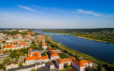 Widok z lotu ptaka na miasto nad Wisłą, z zielonym mostem i zabudowaniami w słoneczny dzień.