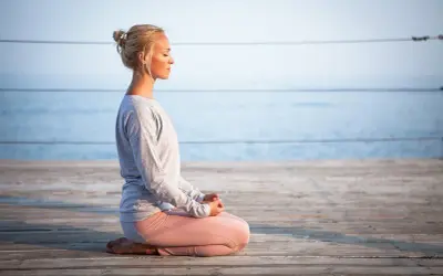 Kvinna med slutna ögon sitter i meditationsställning på en träbrygga vid havet, utövar progressiv avslappning.