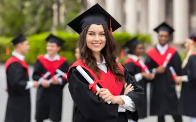 Uśmiechnięta absolwentka z dyplomem w ręku, w tle inni studenci po ang.