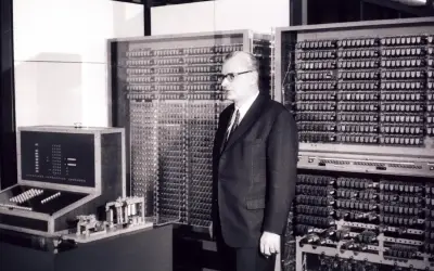 Mężczyzna w garniturze stoi obok ogromnej maszyny z mnóstwem przełączników i lamp. To prawdopodobnie pierwszy komputer na świecie, przełomowa technologia.