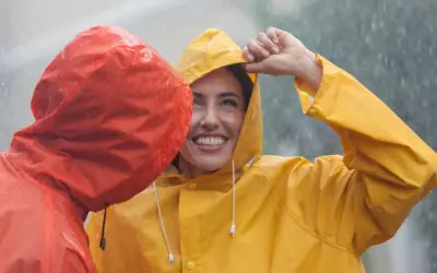 Zwei lachende Personen im Regen, eine in Gelb, die andere in Rot. Die gelbe Regenjacke ist im Fokus.