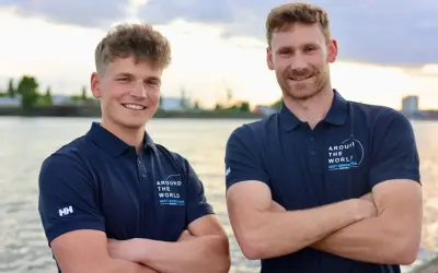Melwin Fink und sein Teamkollege posieren lächelnd am Wasser. Sie tragen marineblaue Poloshirts mit dem Aufdruck "Around The World - Next Generation".