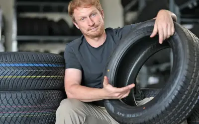 Mann mit roten Haaren hält einen Autoreifen, im Hintergrund Reifenstapel.