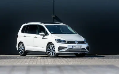 Biały Volkswagen Touran z czarnym dachem i przyciemnianymi szybami, zaparkowany przed ciemną, nowoczesną ścianą.