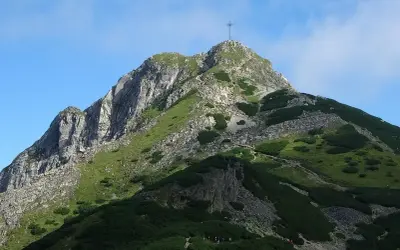 Widok na szczyt Giewontu z krzyżem, zielone zbocza i kamieniste ścieżki. W dole grupa turystów rozpoczyna wejście na Giewont.