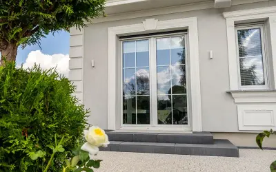 Eleganckie okno bez klamki z białymi szprosami, odbijające błękitne niebo i chmury. Obok zielone krzewy i biała róża.