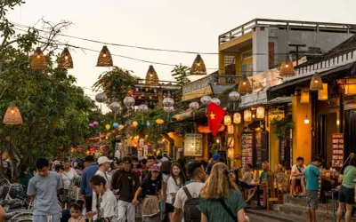 Bunte Lichter und Menschenmassen beleuchten die **Hoi An Altstadt**. Ein vietnamesisches Banner weht über einer belebten Straße voller Geschäfte und Restaurants.