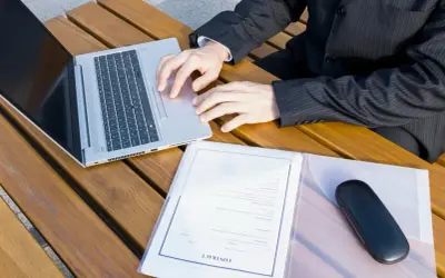 Biznesmen pracuje na laptopie przy drewnianym stole, obok dokumenty z nagłówkiem "CONTRACT". Przygotowuje wpis do księgi wieczystej.