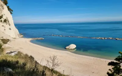 Spiaggia dorata e mare cristallino sulla Riviera del Conero, con una scogliera bianca a picco sull'acqua e un grande masso solitario.