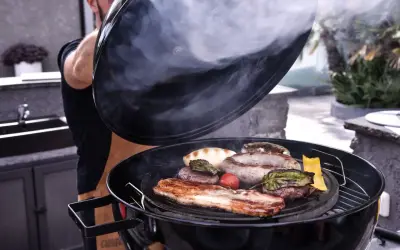 Un homme soulève le couvercle d'un barbecue fumant, révélant des saucisses, des steaks et du bacon. La cuisson lente permet d'attendrir la viande pour un repas savoureux.