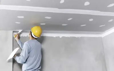 Pracownik w żółtym kasku nakłada masę szpachlową na sufit, przygotowując go do montażu. To kluczowy etap, by wiedzieć, jak przykleić styropian do sufitu.