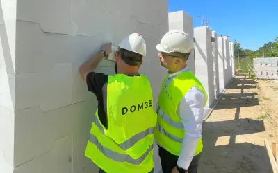 Dwóch mężczyzn w kamizelkach odblaskowych na placu budowy sprawdza postęp prac przy ścianie z białych pustaków.