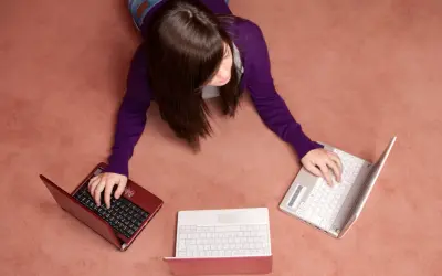 Kobieta leży na dywanie i pisze na trzech laptopach.
