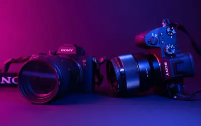 Dwa aparaty Sony, jeden z obiektywem 70-200mm, drugi z 85mm f/1.8. Różne rodzaje aparatów fotograficznych w neonowym świetle.