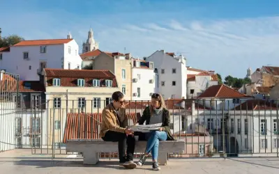 Para planujących tani city break, para siedzi na ławce, studiując mapę na tle urokliwych, białych budynków z czerwonymi dachami.
